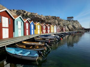 Old, colorfull fishing cabins lining the jetty of Smögen. Several small row boats are tied to the jetty and in the background cliffs are visible.